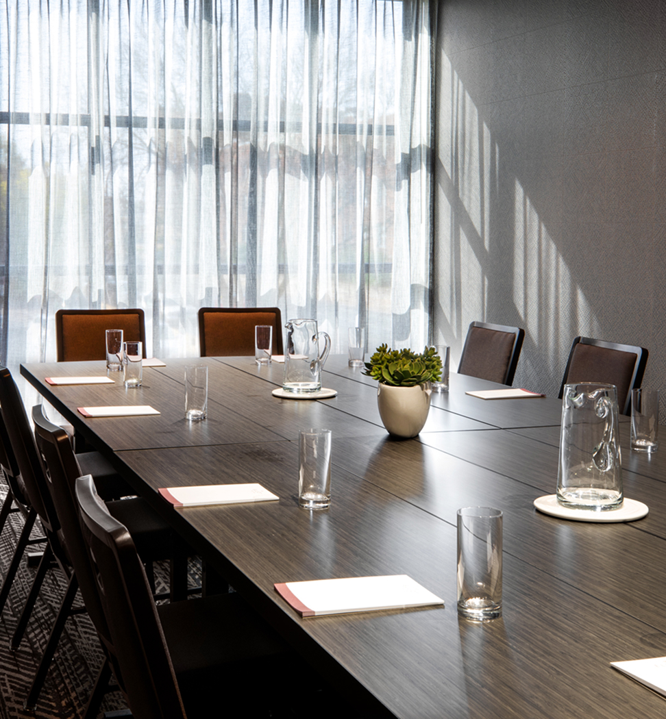 A conference room with a long table set with notepads, glasses, pitchers of water, and a potted plant, with natural light coming through sheer curtains.